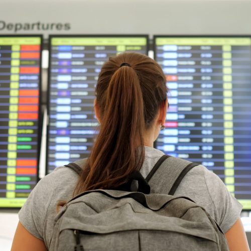 brunette with ponytail and grey backpack wearing t-shirt looks at departures schedule in airport lounge backside view