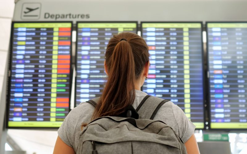 brunette with ponytail and grey backpack wearing t-shirt looks at departures schedule in airport lounge backside view