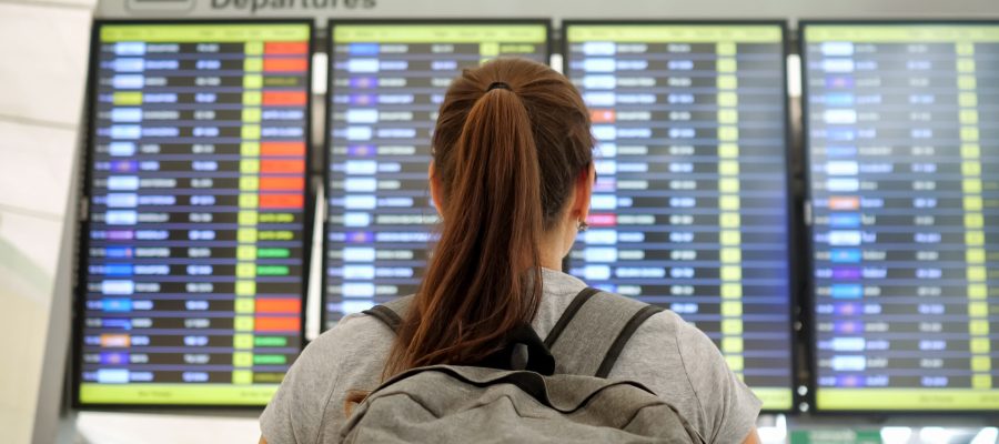 brunette with ponytail and grey backpack wearing t-shirt looks at departures schedule in airport lounge backside view