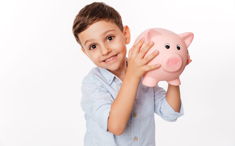Close up portrait of a cheerful cute little kid holding piggy bank and looking at camera isolated over white background