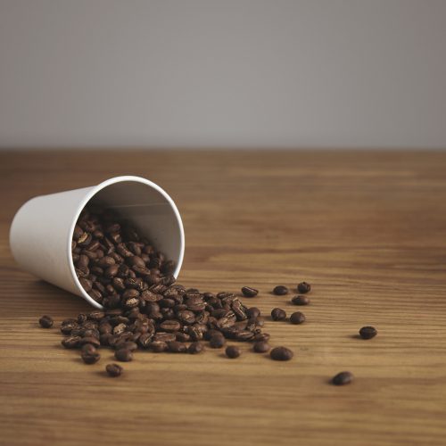 Blank white paper cup with good roasted coffee beans dropped on thick wooden table in cafe shop
