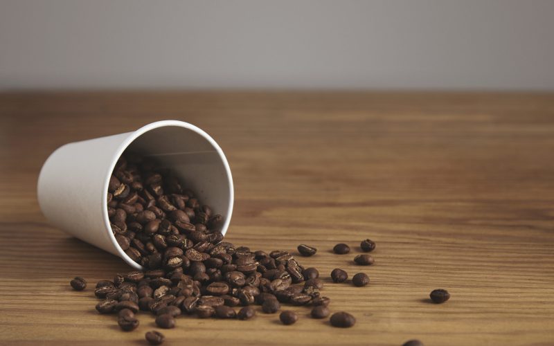 Blank white paper cup with good roasted coffee beans dropped on thick wooden table in cafe shop